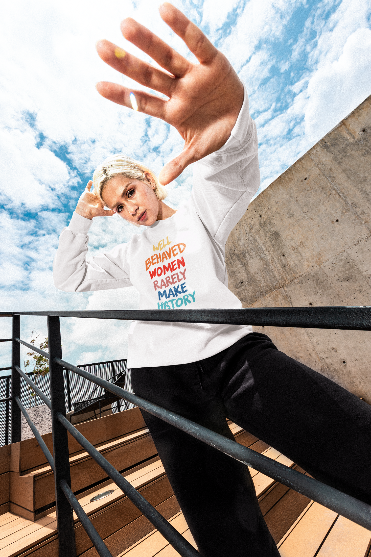 Person wearing a white sweatshirt with colorful text, standing on a rooftop with a blue sky and clouds in the background.