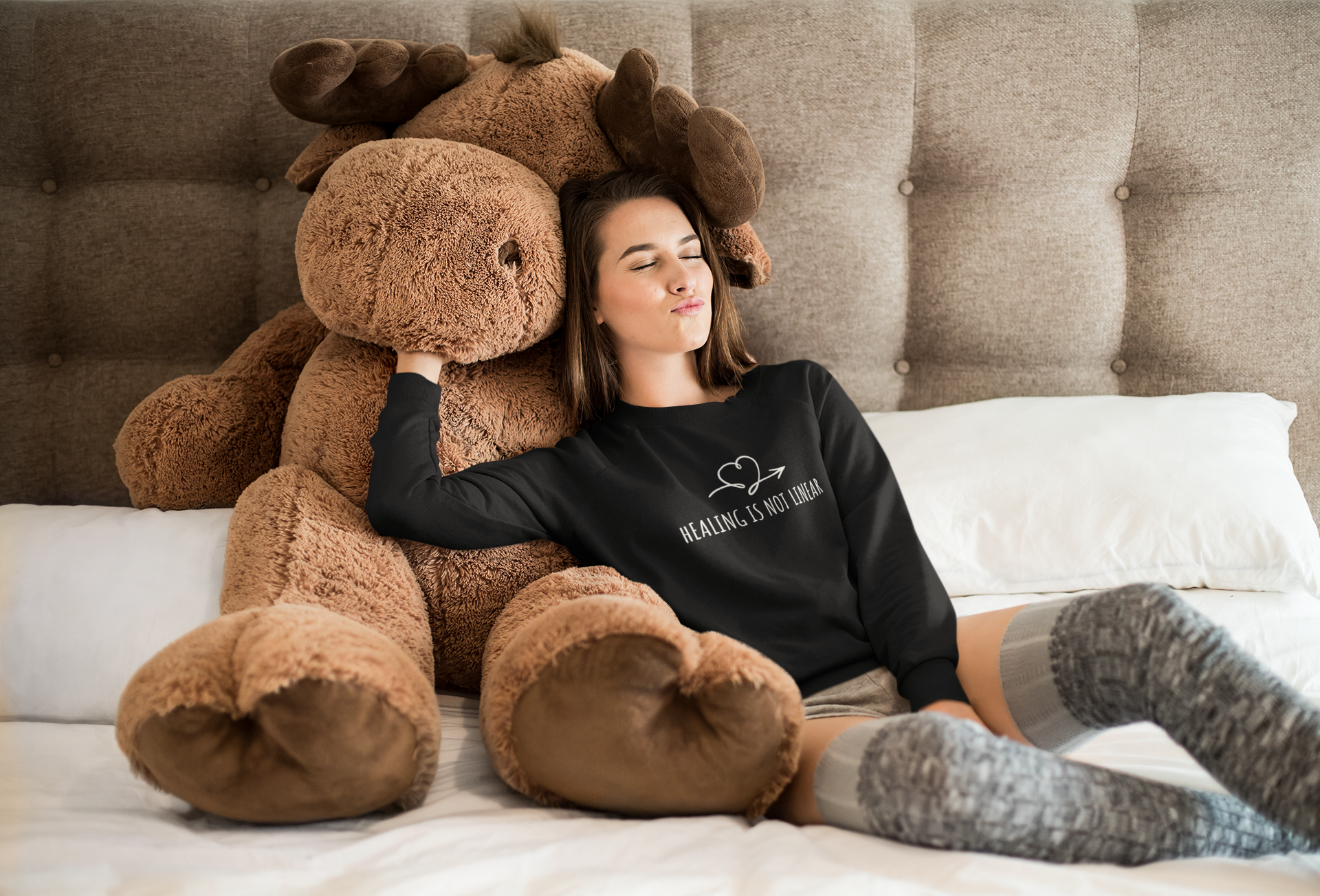 Person sitting on a bed with a large brown teddy bear, wearing a black sweatshirt with a heart design.