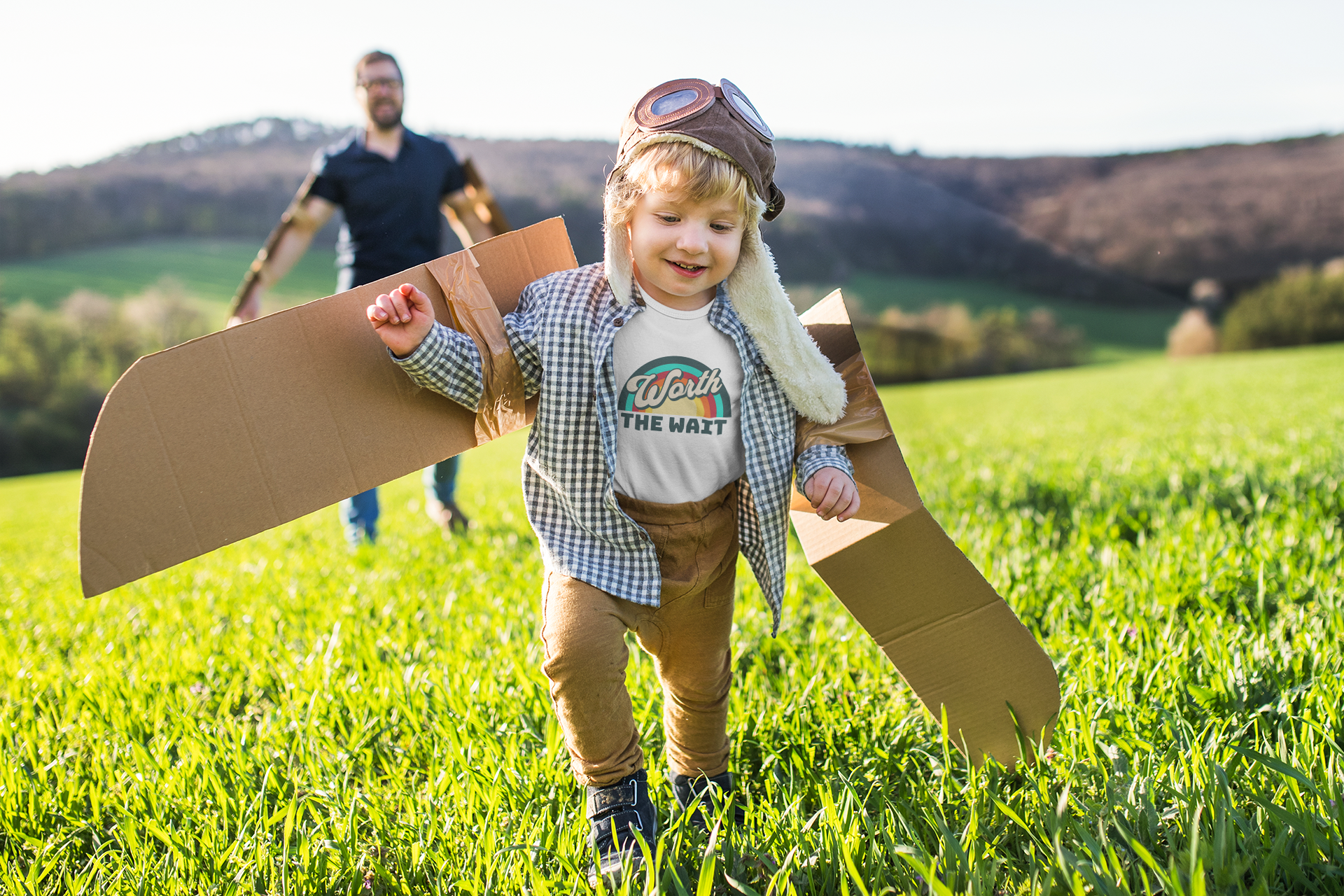 Child wearing revel moki toddler t shirt that says worth the wait, walking with cardboard airplane wings and propeller hat running in a field with a man in the background.