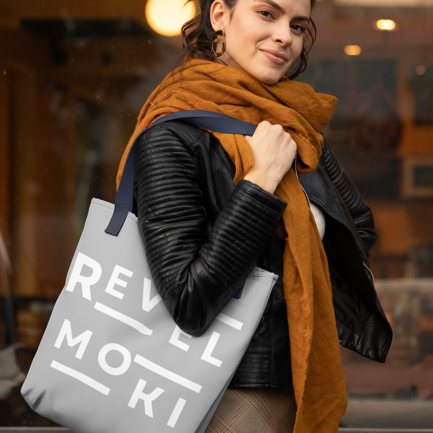 Woman holding a gray tote bag with 'REVEL MOKI' branding, wearing a mustard scarf and black jacket.